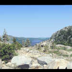 Donner Pass Granite Rock Fields and in the Distance Donner Lake. Donner Pass, CA.  View opens looking east over Donner Lake in the distance. Looking closely, you will see the old rail road tunnel entrance that once carried the first Trans-Continental steam engine powered trains over Donner Pass. This is one of the several routes of the pioneer "California Trail" over the High Sierras. The ox drawn covered wagons had to be moved up a practically vertical granite cliff just beyond those rocks to the east and then had to negotiate around and over the rocks you see everywhere here. In the distance of the east view, you can see the tunnel entrance of the original (first) Trans-Continental RR that came this way. The main Interstate I-80 over the Sierras, from Sacramento to Nevada, several miles North of here, parallels the former Historic California Trail over Donner Pass. It is heart breaking to learn that, among the several different ways over the California High Sierras to the Gold Fields, Donner Pass is not the easiest, or even the shortest route! The emigrants were falsely drawn into this tortured route by literature written (or promoted by) persons motivated by financial gain, certainly not the best interests of those who had to suffer it!  ************************************ (Photo = ~123-2304+7...... ZMM Page = None ...... WayPt = 075 7150ft. My Rand McNally gives 7089 ft)